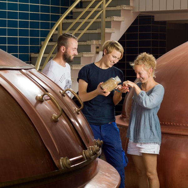 Drei junge Menschen stehen bei einer Führung zwischen Brauereigeräten der Flensburger Brauerei und probieren das Bier.