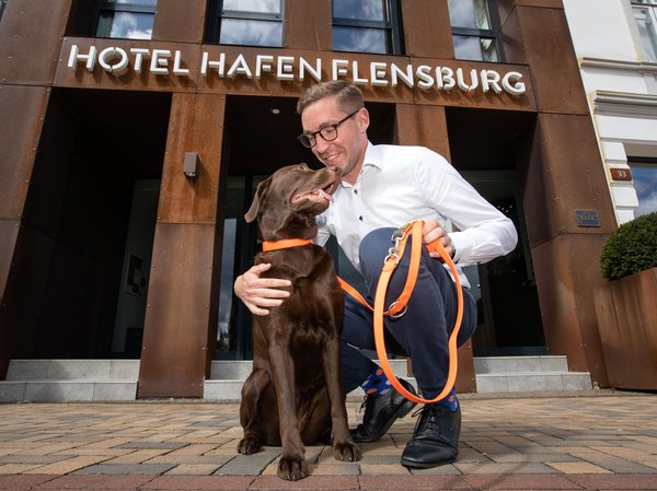 En brun labrador står foran Hotel Hafen Flensburg, hans mester knæler ved siden af ​​ham og strejker hunden.