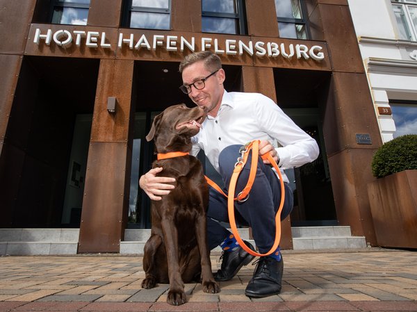 Ein brauner Labrador steht vor dem Hotel Hafen Flensburg, sein Herrchen kniet neben ihm und streichelt den Hund.