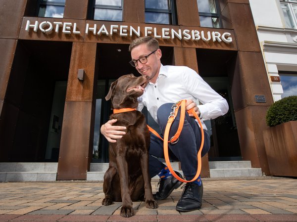 A brown Labrador stands in front of the Hotel Hafen Flensburg, his master kneels beside him and strokes the dog.